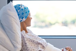 Young woman in bed suffering from cancer. Thoughtful woman battling with tumor looking out of window. Young patient with blue headscarf recovery in hospital on bed.